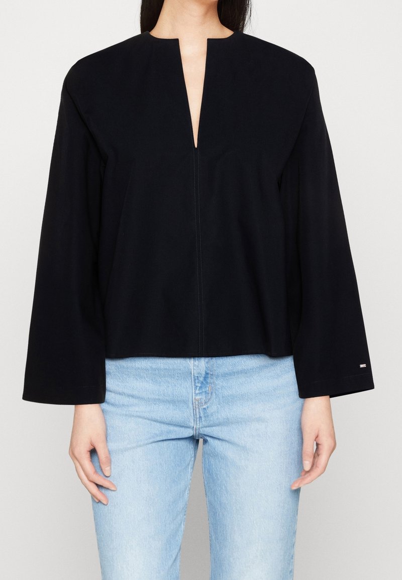 Haut noir avec un décolleté en V profond et des manches larges, fait de tissu lisse. Associé à un jeans bleu clair pour le contraste.