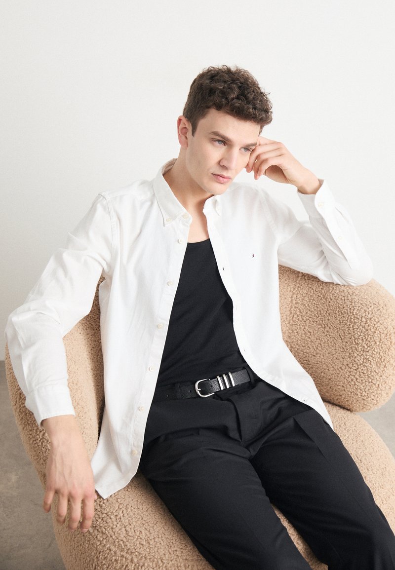 Camicia bianca a maniche lunghe con una texture morbida, indossata sopra un top nero. Cintura nera che aggiunge contrasto. Modello seduto su una sedia beige con una texture particolare.