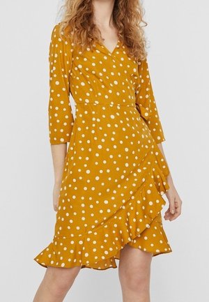 Femme portant une robe portefeuille jaune moutarde à pois blancs avec un ourlet à volants, debout devant un fond clair uni.