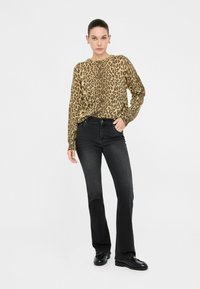 Camicia a maniche lunghe con stampa leopardata in tonalità marrone e beige, abbinata a jeans neri svasati. Modello indossa scarpe nere, mani sui fianchi, sfondo neutro.