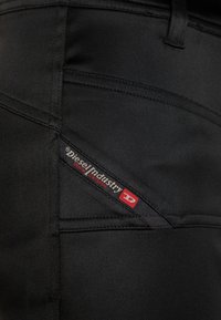 Svarta denimbyxor med en närbild av bakfickan, med en synlig röd och svart varumärkesetikett. Texturerat tyg med detaljerad sömnad.