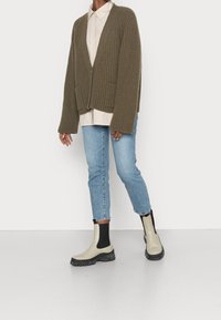 Persona che indossa un cardigan a coste verde oliva, una camicia beige con bottoni, jeans azzurri chiari e stivaletti beige con pannelli elastici neri.