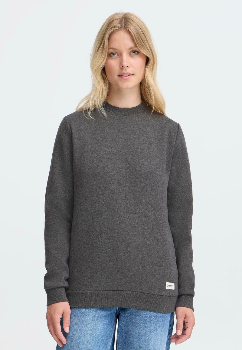 Grå hettegenser med rund hals, ribbestrikkede mansjetter og kant, med et logo-patch nederst til venstre. Glatt, myk stoff.