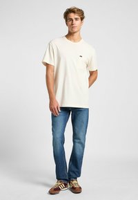 Camiseta de cuello redondo en color beige claro con un bolsillo en el pecho, combinada con jeans ajustados azules y zapatillas marrones con detalles en amarillo.