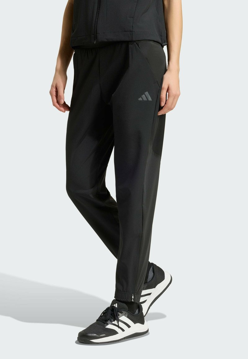 Personne portant un pantalon de sport noir avec le logo Adidas et des baskets noires et blanches, debout avec une jambe croisée sur l'autre.