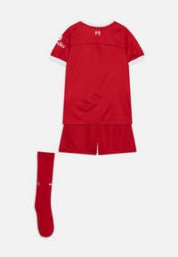 Červený sportovní set skládající se z trička s krátkým rukávem, šortek a ponožky nad kolena. Na tričku jsou bílé akcenty; ponožka má logo.