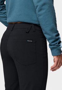 Personne portant un pantalon noir Columbia et un shirt à manches longues couleur sarcelle, montrant la poche arrière et une partie de la main droite.