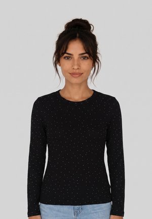 Vrouw met donker haar in een knot, gekleed in een zwart shirt met lange mouwen met kleine witte stippen en lichtblauwe spijkerbroek, staand tegen een grijze achtergrond.