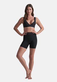 Donna in piedi a piedi nudi, con un reggiseno sportivo nero e pantaloni biker neri a vita alta, mani sui fianchi, sfondo grigio chiaro uniforme.