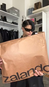 Pessoa com cabelo curto e preto, usando óculos de sol, segurando uma grande sacola de compras castanha da Zalando, em um quarto cheio de roupas e prateleiras.