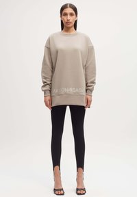 Oversized beige sweatshirt met een ronde halslijn, dropped shoulders en het 'MAISON MISAGA' logo aan de onderkant, gecombineerd met zwarte leggings en hakken.