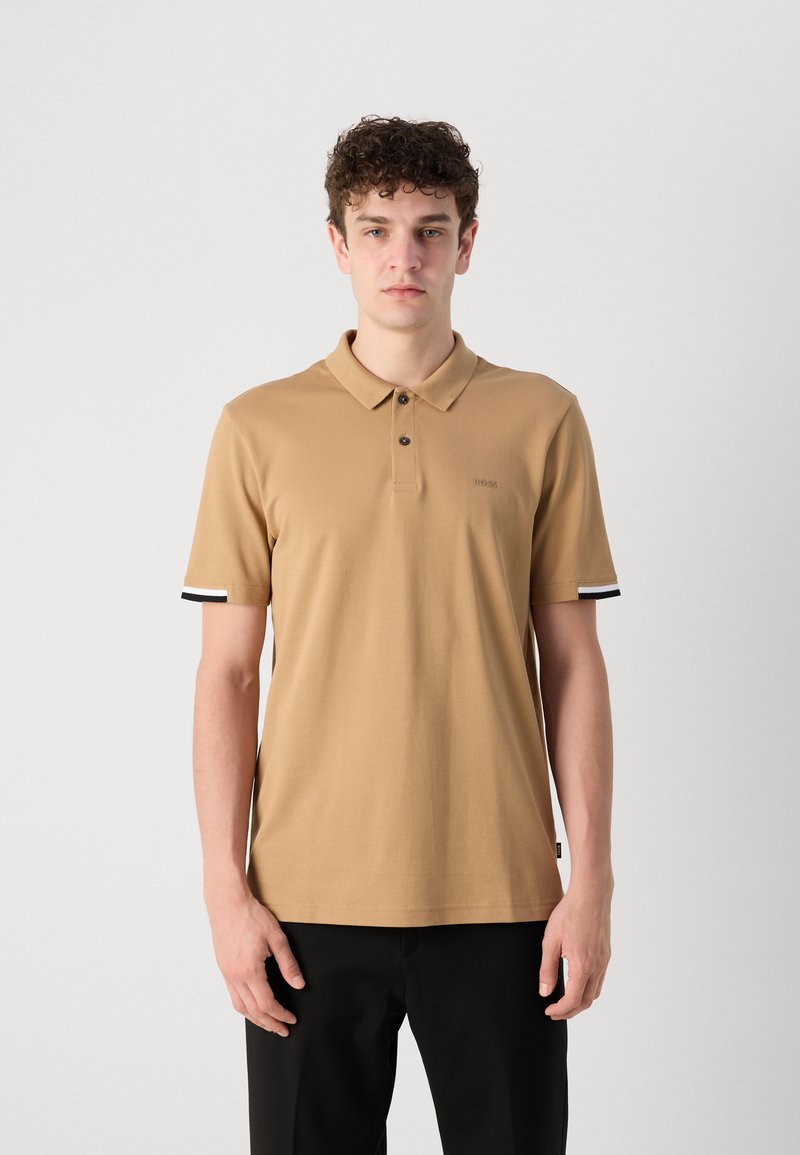Polo shirt beige à manches courtes avec un col, présentant trois boutons et des accents rayés noirs et blancs sur les ourlets des manches.