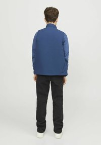 Blå väst med hög krage, parat med en långärmad blå skjorta under, svarta jeans och vita sneakers. Slät tygliknande textur.