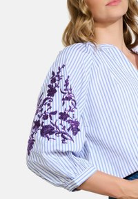 Femme portant un chemisier rayé bleu et blanc avec une broderie florale violette sur des manches bouffantes à trois-quarts.