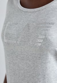 Tricou din bumbac gri cu guler rotund, având inscripțiile "EA7" și "EMPORIO ARMANI" imprimate cu studs metalici argintii.
