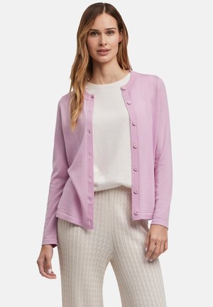 Cardigan rose clair avec un col rond, fermeture à boutons et manches longues, associé à un haut blanc et un pantalon crème texturé.