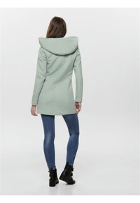 Mintgrüner Mantel mit großer Kapuze, taillierter Form und langen Ärmeln. Getragen mit blauen Jeans und schwarzen Stiefeletten. Glatte Textur.