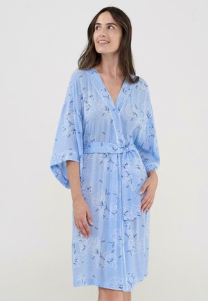 Mujer con un albornoz cruzado de flores azul claro, con mangas sueltas, atado a la cintura, de pie frente a un fondo blanco liso.