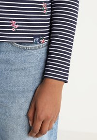 Havsblå och vit randig långärmad skjorta med blomstermönster och en etikett, i kombination med ljusblå denimjeans.