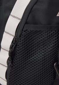 Black backpack made of durable fabric with a mesh section and light gray strap accents. Features a zipper and textured surfaces.