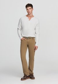 Maglietta henley a maniche lunghe di un grigio chiaro con tre bottoni, abbinata a pantaloni beige a gamba dritta e scarpe marroni con lacci. Design semplice e casual.