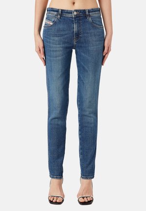 Kobieta w dopasowanych niebieskich dżinsach i czarnych sandałach na obcasie, stojąca ze swobodnie opuszczonymi rękami na tle gładkiej ściany.