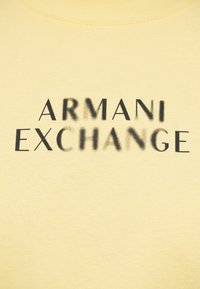 Κίτρινο βαμβακερό μπλουζάκι με το κείμενο "ARMANI EXCHANGE" σε έντονη μαύρη γραμματοσειρά, με ελαφρύ σκιάδασμα στα γράμματα.