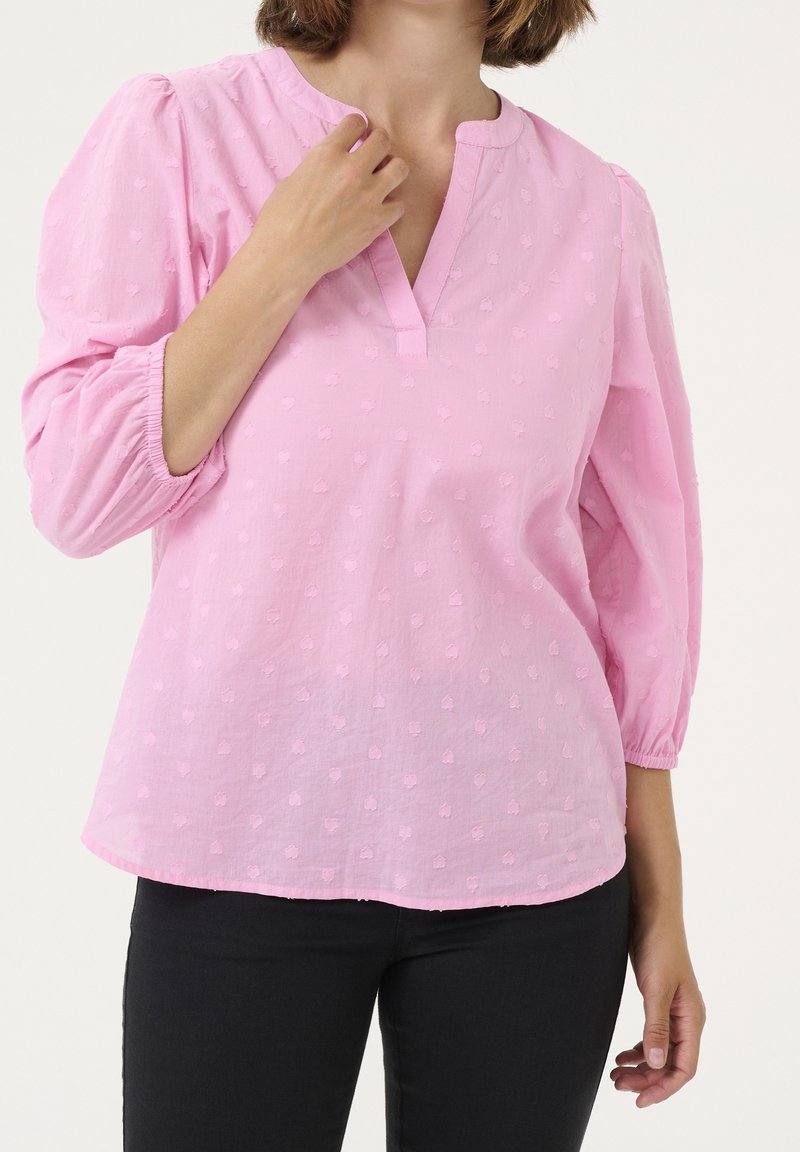 Blouse en coton rose avec un décolleté en V, des manches bouffantes et des pois texturés. Associée à un pantalon noir, mettant en avant une coupe décontractée et un design simple.