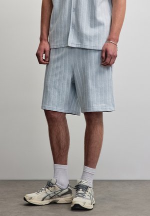 Mann trägt hellblaue, strukturierte Shorts, passendes kurzärmliges Hemd, weiße Socken und weiße und graue Sneakers, steht auf grauem Boden.
