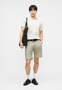 Νέος άνδρας που φοράει μπεζ μπλουζάκι και σορτς, μαύρη ζώνη και αθλητικά παπούτσια, κουβαλώντας μια μεγάλη μαύρη τσάντα ώμου.