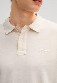 Beige poloshirt van textiel met structuur; voorzien van een klassieke kraag, een tweeknoopsluiting en een reliëf "JOOP! Jeans" logo op de borst.