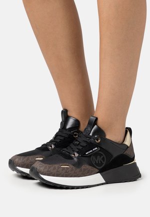 Czarne i brązowe sneakersy Michael Kors ze złotymi przelotkami i białymi środkowymi podeszwami, noszone na boso na jednolitym tle.