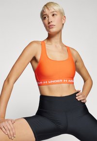 Lys oransjefarget sports-BH med et bredt hvitt bånd som har «UNDER ARMOUR»-tekst, sammen med høytlivs svarte shorts. Glatt stofftekstur.