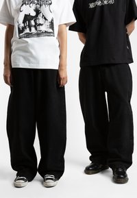 Two individuals wear oversized black pants. One wears a white graphic t-shirt, the other a black shirt with text. Footwear includes sneakers and black boots.