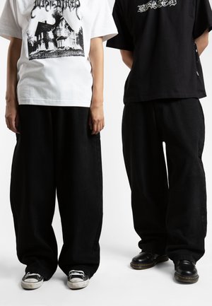 Two individuals wear oversized black pants. One wears a white graphic t-shirt, the other a black shirt with text. Footwear includes sneakers and black boots.
