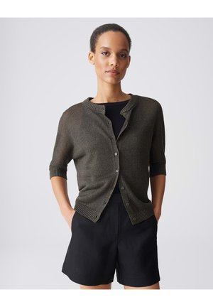 Frau mit kurzen Haaren, die eine dunkelgrüne, geknöpfte Strickjacke über einem schwarzen Top und schwarzen Shorts trägt, steht mit den Händen in den Taschen vor einem schlichten Hintergrund.