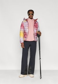 Πανωφόρι με επένδυση και χρωματικές μπλοκ σε ροζ, μωβ και κίτρινες αποχρώσεις· συνδυασμένο με ροζ πουλόβερ, σκούρο παντελόνι και ανοιχτόχρωμα παπούτσια· κρατώντας μπαστούνια πεζοπορίας.