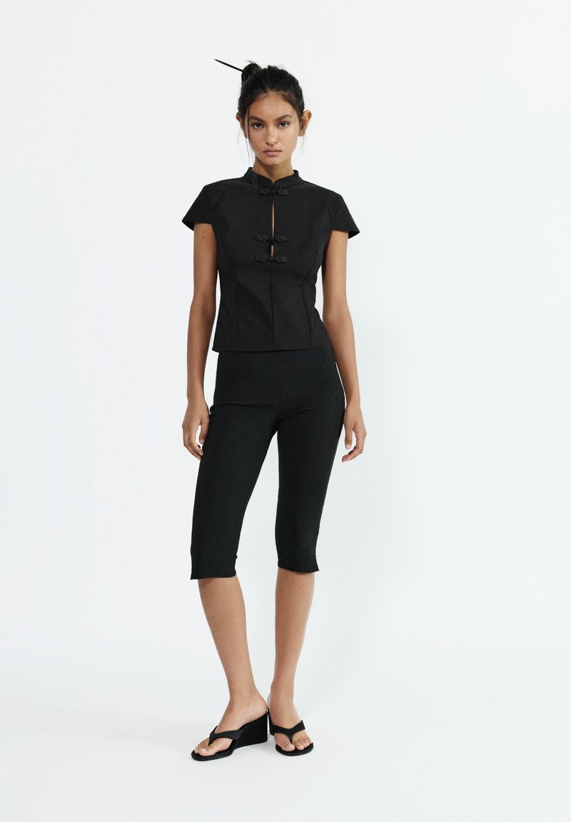 Femme debout portant un haut noir ajusté à manches courtes avec des fermetures à grenouilles, des leggings noirs courts et des sandales compensées noires sur fond blanc.
