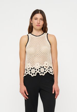Femme debout face à l'observateur, portant un top beige en crochet sans manches avec un liseré noir et un ourlet fleuri, assorti à un pantalon noir sur fond uni.
