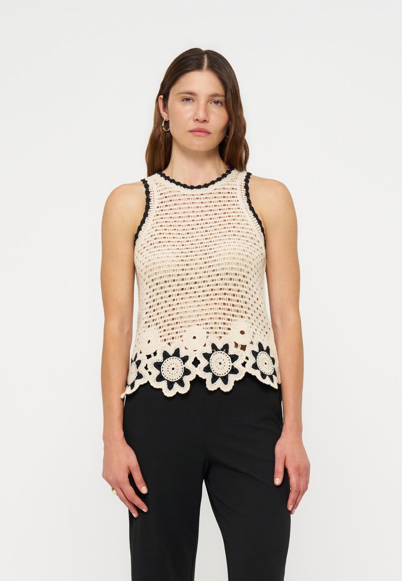 Femme debout face à l'observateur, portant un top beige en crochet sans manches avec un liseré noir et un ourlet fleuri, assorti à un pantalon noir sur fond uni.