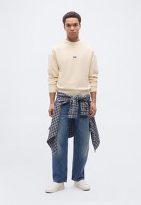 Jeune homme debout, vêtu d'un sweat-shirt crème, d'un jean bleu, de baskets blanches et d'une chemise grise à carreaux nouée autour de la taille, sur un fond blanc.