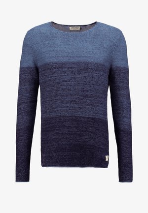 Jeune homme portant un pull bleu dégradé, un jean foncé, des baskets blanches et un sac à dos marron, debout les mains près des poches du jean.