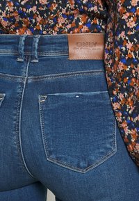 Dunkelblaue Denim-Jeans mit einer strukturierten Oberfläche. Verfügt über ein beige Lederpatch am Bund und zwei Gesäßtaschen mit kontrastierenden Nähten.