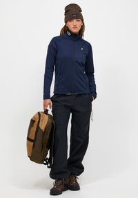 Giacca a zip navy, pantaloni neri, berretto marrone, tiene uno zaino marrone e oliva. Materiali testurizzati, design resistente alle intemperie. Stivali da trekking.