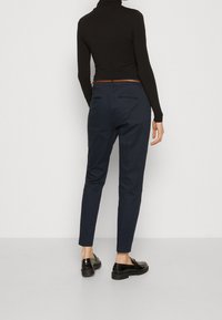 Navy tapered trousers with a smooth texture, back pockets, and a subtle belt detail, paired with black shoes on a neutral background.