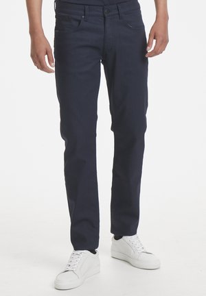 Jean en denim bleu foncé à coupe slim, cinq poches et une texture subtile. Associé à des baskets blanches à semelle basse.