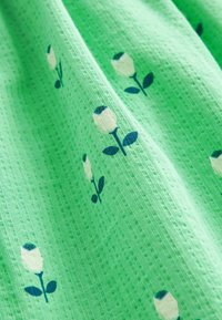 Vihreä kangas, jossa on teksturoitu kuvio, joka esittää pieniä valkoisia ruusujemerkkejä sinisillä korostuksilla ja tummanvihreillä lehdillä.