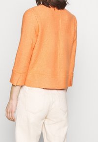 Veste orange texturée courte avec manches trois-quarts et poignets légèrement évasés, portée par-dessus un pantalon de couleur claire. Vue de dos.