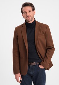 Brun ullblazer med notch-lapel, bärs över en svart polotröja och kombineras med mörka denimjeans. Lätt textur och skräddarsydd passform.