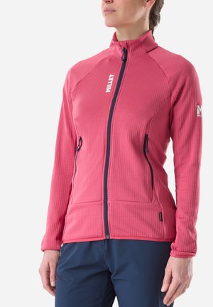 Woman wearing a fitted pink zip-up jacket with two zipped pockets and dark blue pants, standing against a plain background.